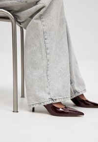 Burgundijas lakoti ādas augstpapēžu kurpes ar smailo purngalu, kas apvienotas ar gaiši pelēkām platām džinsu biksēm, sēžot blakus metāliskai krēsla.