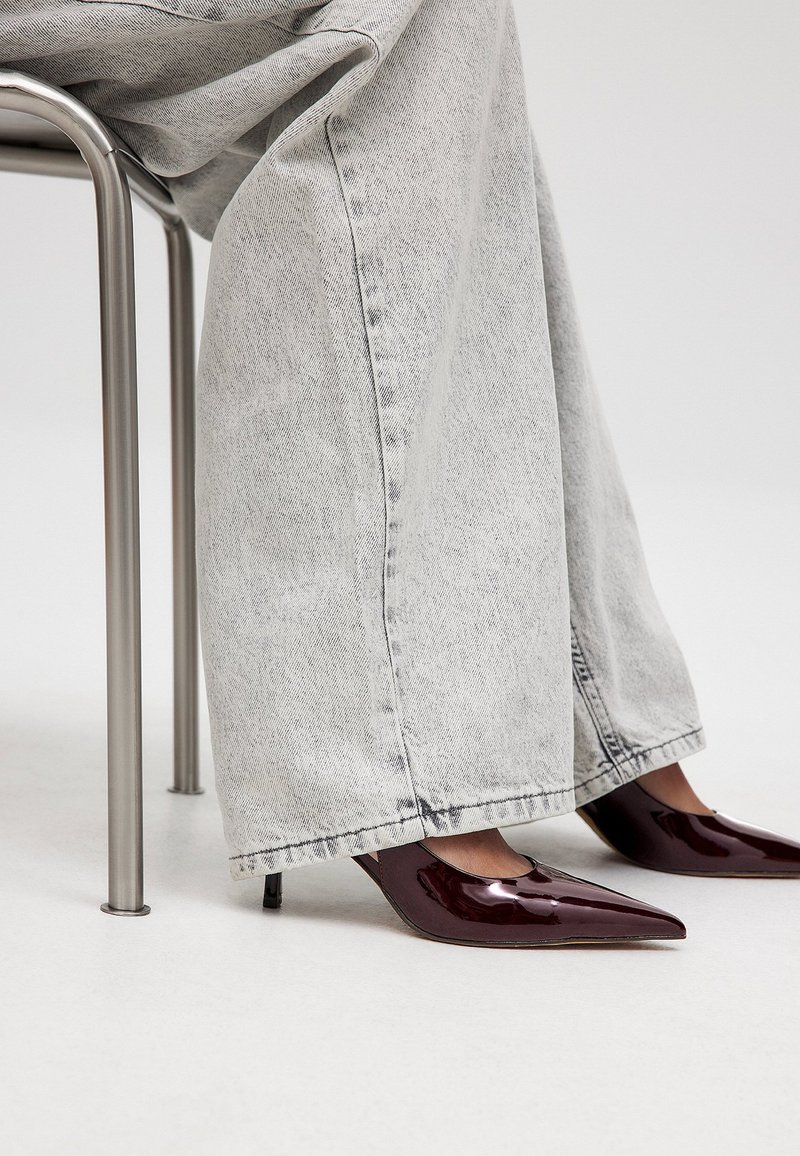 Burgundijas lakoti ādas augstpapēžu kurpes ar smailo purngalu, kas apvienotas ar gaiši pelēkām platām džinsu biksēm, sēžot blakus metāliskai krēsla.