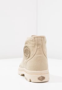 Béžová plátěná kotníkova obuv s texturovaným povrchem, kruhovým logem na patě a robustní gumovou podrážkou se zigzagovým vzorem.