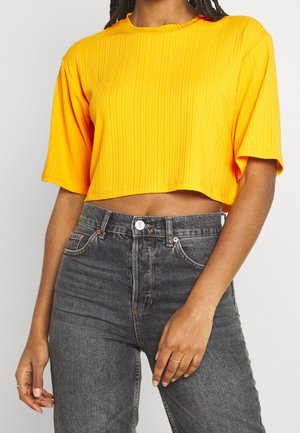 Mujer que lleva una camiseta corta de canalé de color amarillo brillante con mangas cortas, combinada con vaqueros negros lavados de talle alto.