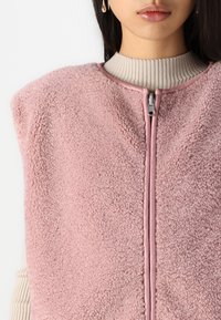 Vesta roz din fleece cu o suprafață texturată și un guler înalt. Prezintă un fermoar frontal și un guler interior cu nervuri de culoare deschisă contrastantă.