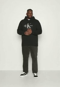 Moletom preto com o logótipo "Calvin Klein Jeans", combinado com calças cinza-escuras e ténis preto e branco, de pé contra um fundo simples.