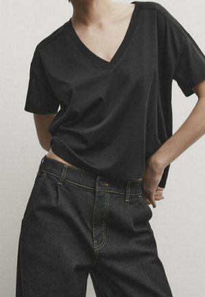 Person iført en løs sort V-hals T-shirt, der er let stoppet ind i mørkeblå, højtaljede denimjeans, med hænderne hvilende på hofterne.