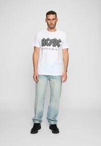 Hvid bomulds t-shirt med sort grafik "AC/DC" og teksten "BACK IN BLACK". Iført med lyseblå jeans og sorte sneakers, stående oprejst.
