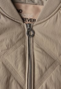 Veste beige matelassée avec une fermeture éclair centrale argentée équipée d'un bouton-poussoir circulaire et un texte visible à l'intérieur du col indiquant "PARADISE FOREVER".