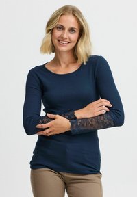 Námornícky modrý dlhý rukáv s čipkovými akcentmi na manžetách, s rebrovanou textúrou a okrúhlym výstrihom. Model má ruky prekrížené pred sebou.