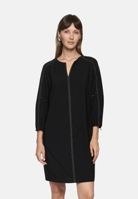 Schwarzes Kleid mit V-Ausschnitt, dreiviertellangen Ballonärmeln, kontrastierenden Nähten und einem Reißverschluss vorne. Glatte Stofftextur.