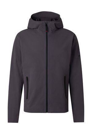 Blonde Person trägt dunkelgraue Kapuzenjacke, passende Hose, schwarze Stiefel und übergroße schwarze Sonnenbrille, steht vor weißem Hintergrund.