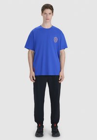 Jeune homme aux cheveux courts bruns portant un T-shirt bleu oversize avec texte, un pantalon de jogging noir et des baskets noires, debout sur un fond gris.