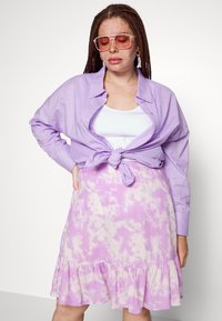 Camisa lilás de botão amarrada na cintura, usada sobre uma blusa branca, combinada com uma saia lilás em camadas com um padrão tie-dye.