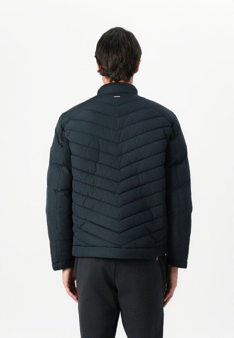 Ciemnoniebieska kurtka puchowa z wzorem w zygzaki, wysokim kołnierzem i małym logo z tyłu. Posiada długie, lekko zwężane rękawy.