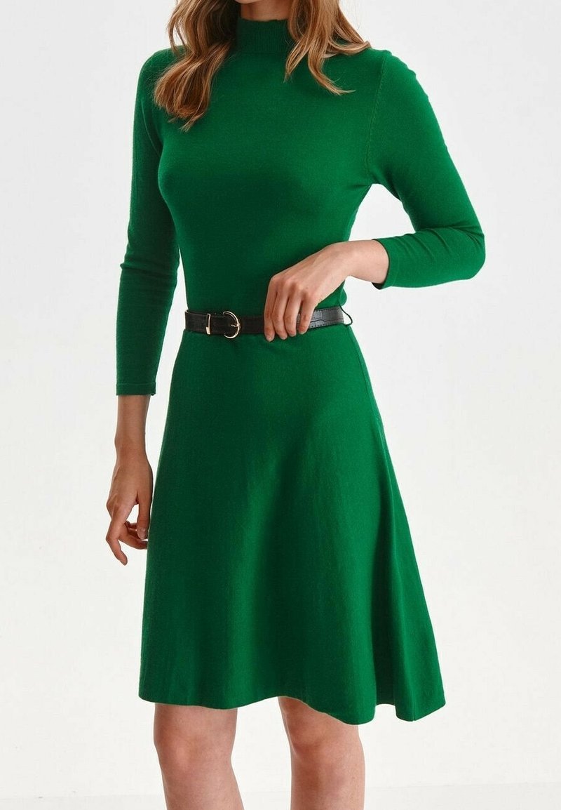 Grön, stickad klänning med figursydd överdel, långa ärmar och utsvängd kjol; har ett svart bälte med en rund spänne i midjan.