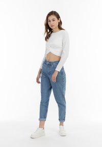 Top cropped bianco a maniche lunghe con dettaglio nodo, abbinato a jeans high-waisted blu chiaro e sneakers bianche. Design semplice, vestibilità casual.