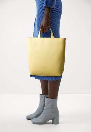 Person in blue ribbed dress holding yellow tote bag, wearing light gray ankle boots with block heels on white background.
