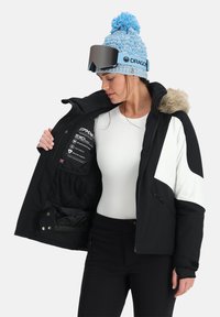 Giacca nera isolata con dettagli bianchi, cappuccio con bordo in pelliccia, zip frontale e numerose tasche interne. Indossata sopra una maglietta bianca.