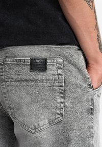 Des jeans en denim gris présentent une poche arrière avec une étiquette noire indiquant "Ombre". Le tissu a une finition texturée et une apparence usée.