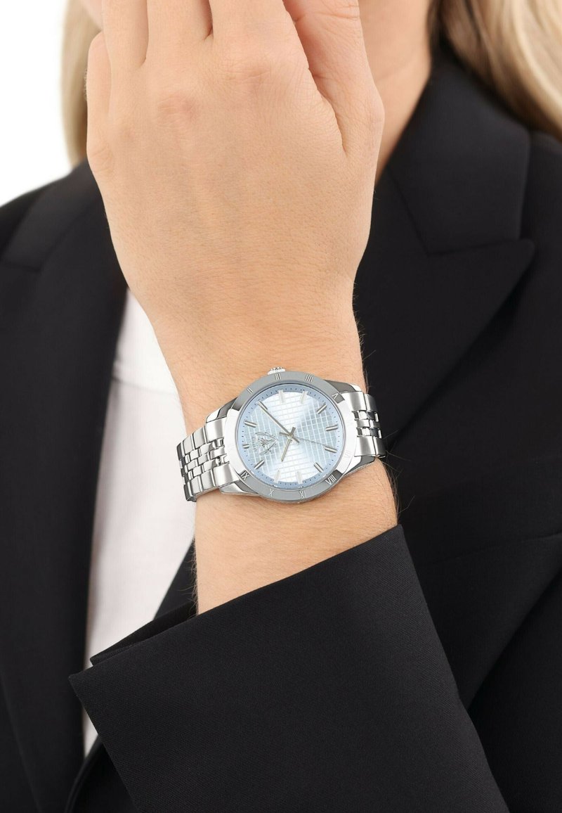 Armbanduhr mit silbernem Metallarmband und hellblauem gemusterten Zifferblatt, getragen am linken Handgelenk einer Person in schwarzem Blazer.