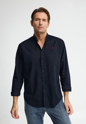 Uomo in piedi contro uno sfondo bianco che indossa una camicia nera a maniche lunghe abbottonata e jeans blu con le maniche arrotolate.