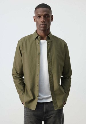 Hombre vistiendo una camisa de botones verde oliva sobre una camiseta blanca y vaqueros oscuros, de pie con las manos en los bolsillos contra un fondo liso.