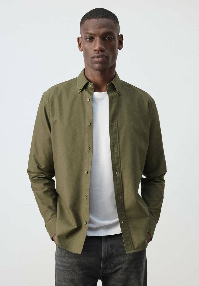 Homme portant une chemise vert olive boutonnée sur un t-shirt blanc et un jean foncé, debout les mains dans les poches devant un fond uni.