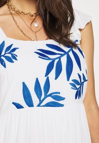 Vrouw in een witte jurk met blauw geborduurde bladpatronen en gelaagde gouden kettingen met een ronde hanger.