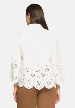 Hvid bluse med klokkeærmer, der har bølget kant og blomstrede øjenhulsudskæringer. Glat stof med løs pasform, kombineret med brune bukser.