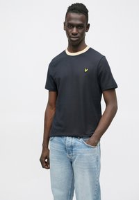 Fekete rövid ujjú póló kerek nyakkivágással és bézs szegéllyel, apró sárga logóval a mellrészen. Világoskék farmernadrággal viselve.