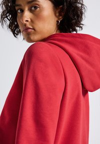 Femme aux cheveux bouclés portant un sweat à capuche rouge, se retournant sur son épaule devant un fond blanc.
