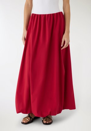 Donna che indossa una lunga gonna rossa, ampia con orlo arricciato, abbinata a sandali piatti neri con dettagli dorati.