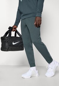 Donker teal joggingsbroeken met geribbelde boorden, gecombineerd met witte Nike sneakers. Een zwarte Nike sporttas met een wit logo wordt in één hand vastgehouden.