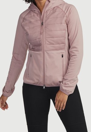 Henkilö, jolla on vaalean liilanvärinen, hupullinen, tikattu vetoketjullinen takki ja mustat housut, seisomassa rentoina käsin sivuilla.