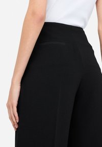 Pantalones negros a medida con una textura de tela suave, un bolsillo trasero único con un ribete sutil y un diseño de pierna recta.