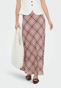 Jupe longue à carreaux beige avec des nuances bordeaux et crème, associée à un gilet blanc ajusté et un sac fourre-tout blanc texturé. Chaussures en cuir marron.