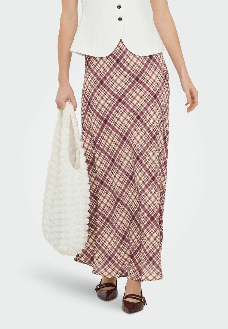 Jupe longue à carreaux beige avec des nuances bordeaux et crème, associée à un gilet blanc ajusté et un sac fourre-tout blanc texturé. Chaussures en cuir marron.