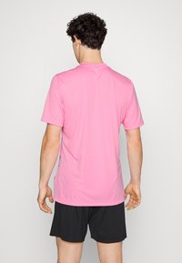 Maglietta sportiva a maniche corte di colore rosa chiaro, realizzata in materiale liscio ed elastico, abbinata a pantaloncini neri, vista da dietro.