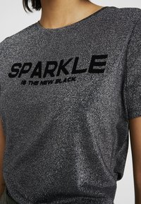 Černé triko s glittry s výrazným, měkkým textem, který říká "JISKRA JE NOVÁ ČERNÁ." Elegantní design s volným střihem a jemnou Texturou.