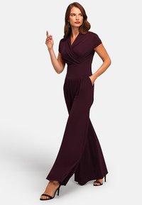 Dark purple jumpsuit with a wrap design, short sleeves, wide legs, and side pockets. Worn with black strappy heels. Smooth fabric texture.