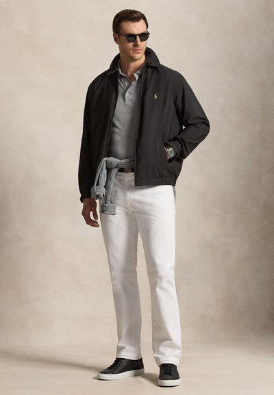 Férfi fekete dzsekiben, szürke pólóban, fehér nadrágban, fekete sportcipőben és napszemüvegben, szürke pulóvert az ágyékán megkötve, bézs háttér előtt pózol.