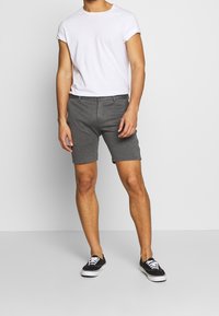 Pantaloni corti grigi realizzati in materiale morbido, con vestibilità slim e tasche frontali, abbinati a una maglietta bianca con maniche arrotolate e sneakers nere.