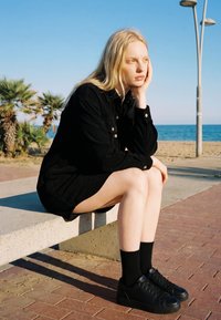 Junge Frau in schwarzem Kleid und Schuhen sitzt auf einer Bank am Strand, stützt ihren Kopf auf die Hand und schaut nachdenklich.