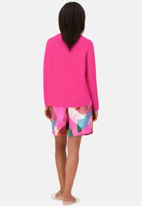 Fuchsia gestrickter Pullover kombiniert mit farbenfrohen, geometrisch gemusterten Shorts. Das Model trägt leichte, flache Schuhe und ist von hinten zu sehen.