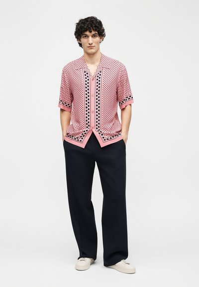 Νέος άντρας με σγουρά μαλλιά που φοράει ροζ μπλούζα με κουμπιά και σύντομο μανίκι με σχέδια, μαύρο φαρδύ παντελόνι και λευκά αθλητικά παπούτσια, με τα χέρια στις τσέπες.