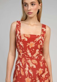 Robe en tissu rouge avec un décolleté carré, ornée de motifs floraux et de paisley blancs et orange, et dotée de larges bretelles.