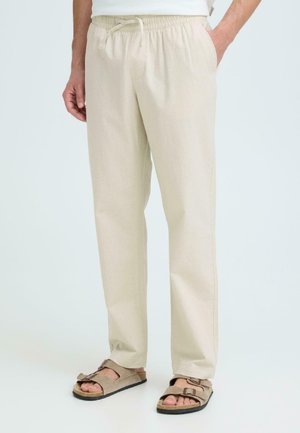 Mann iført beige bukser med avslappet passform, elastisk midje og snor, kombinert med brune sandaler med åpne tær mot en lys bakgrunn.