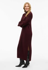 Abito lungo a coste di colore borgogna con collo alto, dotato di spacchi laterali. Abbinato a stivali neri con punta appuntita, che sottolineano una silhouette elegante.