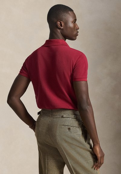 Άνδρας με πλάγια όψη που φοράει κόκκινο πόλο με κοντό μανίκι και μπεζ καρό παντελόνι, μπροστά σε απλό μπεζ φόντο.