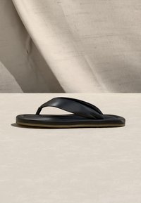 Černá podložená žabková sandála s širokými pásky ležící na světle béžovém povrchu na pozadí béžové látky.