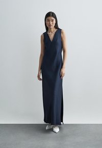 Γυναίκα που φοράει μακρύ μπλε φόρεμα χωρίς μανίκια με λευκά μυτερά παπούτσια και πέρλες στο λαιμό, στέκεται σε γκρι πάτωμα.