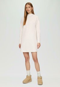 Helles beige gestricktes Kleid mit langen Ärmeln und hohem Kragen, das über eine Nahtdetails an der Vorderseite verfügt. Kombiniert mit hellen Socken und beigen Stiefeletten.