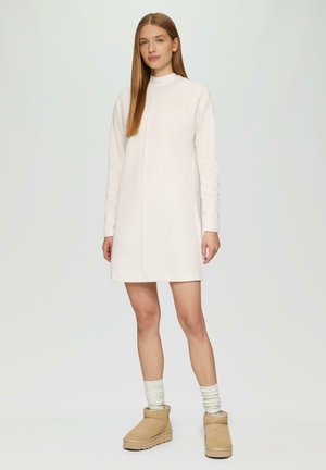 Robe tricotée beige clair à manches longues et col montant, avec un détail de couture à l'avant. Associée à des bottes à la cheville beige et de légères chaussettes.